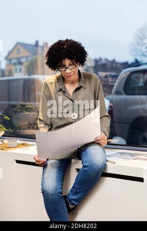Geschäftsfrau, die auf Blaupause schaut, während sie auf der Fensterbank sitzt Stockfoto