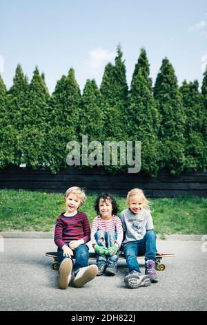 Portrait von glücklichen Kindern, die auf dem Skateboard gegen Bäume sitzen Stockfoto