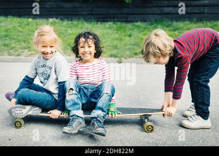 Portrait von glücklichen Kindern auf Skateboard sitzen, während Freund ziehen Es auf dem Hof Stockfoto