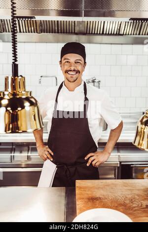 Porträt eines glücklichen männlichen Küchenchefs, der mit den Händen auf den Hüften steht In der kommerziellen Küche Stockfoto
