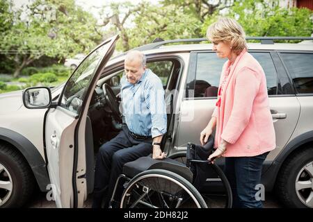 Mann, der aus dem Auto aussteigt, auf dem Rollstuhl, der von einer Frau gehalten wird Stockfoto