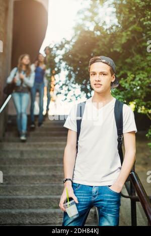 Portrait von selbstbewussten Teenager hält Einweg-Glas während des Stehens auf Schritte mit Freunden im Hintergrund Stockfoto