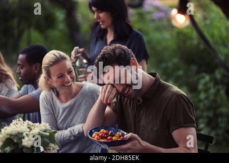 Glückliche Frau, die den Mann ansieht, der die Augen reibt, während sie Tomate hält Schüssel bei der Dinner-Party im Hof Stockfoto