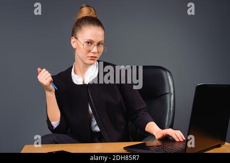 Seriöse, junge und selbstbewusste Geschäftsfrau sitzt mit Brille am Computer und posiert auf grauem Hintergrund. Stockfoto