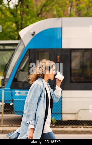 Seitenansicht einer jungen Frau, die Kaffee an der Straßenbahnhaltestelle trinkt Stockfoto