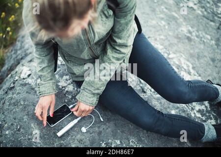 Die Frau, die ein tragbares Ladegerät mit einem intelligenten Gerät verbindet, ist aus einem hohen Blickwinkel zu sehen Telefon auf Stein Stockfoto