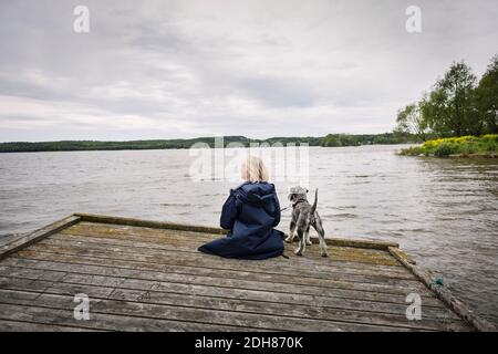 Rückansicht der älteren Frau und des Hundes am Pier see gegen bewölkten Himmel Stockfoto