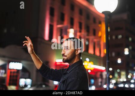 Seitenansicht eines jungen Mannes, der ein Taxi auf der Stadtstraße hagelt Nachts Stockfoto