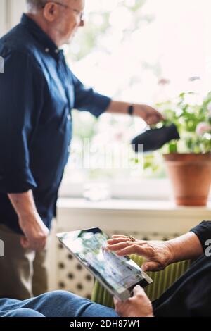 Zugeschnittenes Bild von älteren Frau mit digitalen Tablet, während Mann Bewässerung Blume Pflanze Stockfoto