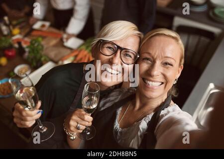 Fröhliche reife Frauen halten Champagnerflöten, während sie Selfie in Küche Stockfoto