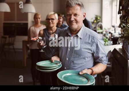 Portrait von reifen Mann hält Platten mit Freunden im Hintergrund Zu Hause Stockfoto