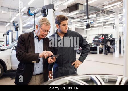 Kunde, der ein Mobiltelefon verwendet, während er mit einem Mechaniker im Auto steht Reparaturwerkstatt Stockfoto