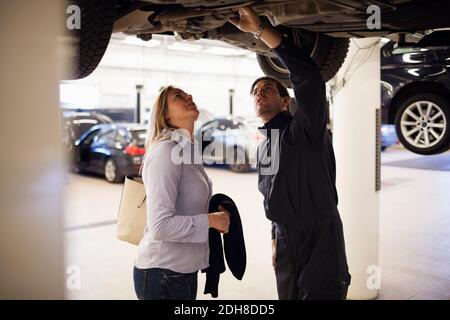 Männlicher Experte mit Kunde, der unter dem Auto in der Werkstatt steht Stockfoto