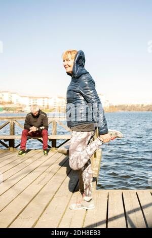 Ältere Frau streckt das Bein, während der Mann auf der Bank sitzt pier gegen klaren Himmel Stockfoto