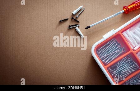 Draufsicht auf Kunststoffstopfen und Schraube in weiß-orange Kunststoffbox. Schraubendreher, Wandstecker und schwarze Schrauben auf brauner Papierkartonstruktur. Satz Schrauben. Stockfoto