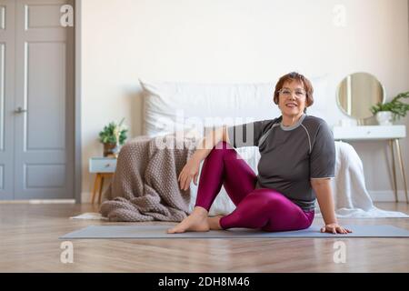 Ältere Frau, die vor einem Pilates-Training auf dem Boden sitzt. Helathy Lifestyle-Konzept. Sie schaut auf die Kamera und lächelt. Stockfoto
