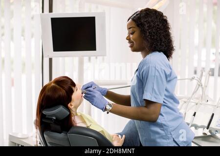 Hübsche afrikanische Zahnärztin trägt Handschuhe, Durchführung zahnärztliche Untersuchung und Behandlung von Patienten Zähne. Junge Frau liegt im Stuhl, bekommen zahnärztliche Stockfoto