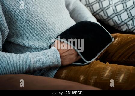 Nahaufnahme eines jungen kaukasischen Mannes mit einem elektrischen heißen Wasserbeutel, um seine Hände zu wärmen, während er sitzt Ein bequemes braunes Sofa Stockfoto