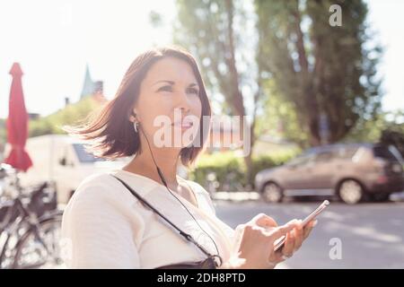 Durchdachte Geschäftsfrau, die wegschaut, während sie Musik über das Smartphone hört An sonnigen Tag Stockfoto