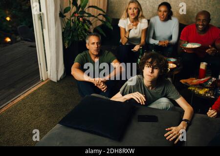 Männlicher Teenager auf dem Sofa beim Fernsehen während der Sportveranstaltung mit Familie bei Nacht Stockfoto