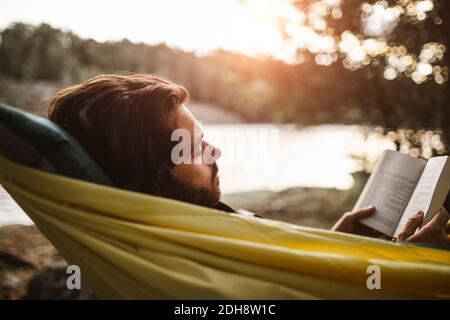 Betrachtend Mann Buch beim Liegen über Hängematte im Wald lesend Stockfoto