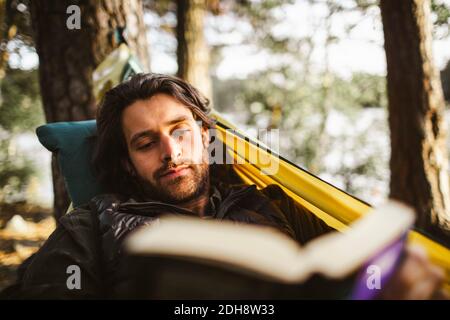 Junger Mann liest Buch, während er in der Hängematte im Wald liegt Stockfoto