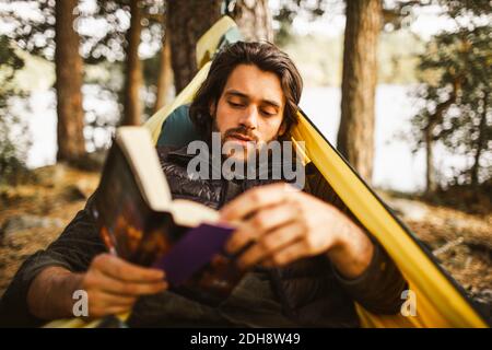 Junger Mann, der Buch liest, während er in der Hängematte liegt Wald Stockfoto