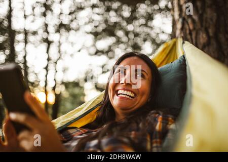 Frau, die Smartphone benutzt, während sie über Hängematte im Wald liegt Stockfoto