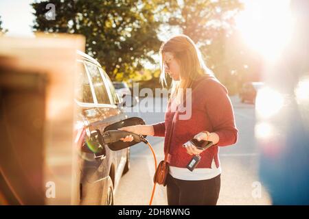 Frau hält Smartphone, während das Elektroauto an der Station geladen wird An sonnigen Tag Stockfoto