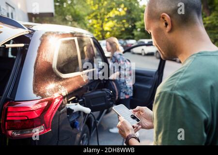 Mann mit Smartphone mit Frau, die mit dem Auto im Hintergrund steht Stockfoto