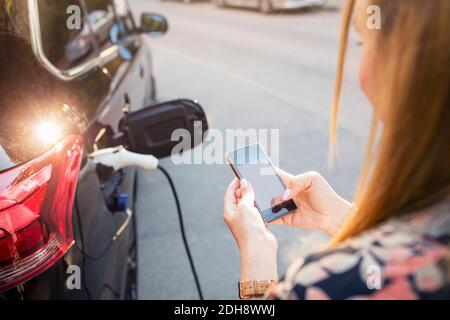 Frau, die ein Mobiltelefon benutzt, während sie im Stehen steht und ein Elektroauto lädt Stockfoto