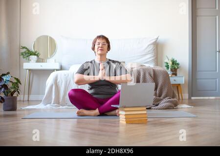 Eine ältere Frau meditiert in Lotushaltung zu Hause vor einem Laptop-Monitor. Das Konzept eines gesunden und aktiven Lebensstils im Alter. Stockfoto