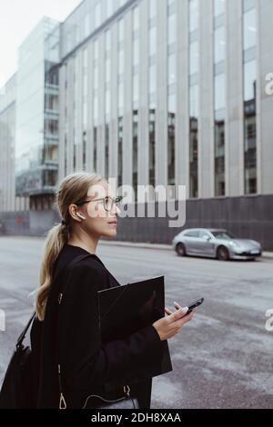 Seitenansicht einer Geschäftsfrau mit in-Ear-Kopfhörern, die die Datei in der Hand halten Stadt Stockfoto