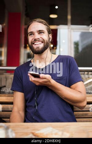 Lächelnder Mann hört Musik über Smartphone, während er sitzt Straßencafé Stockfoto