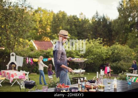 Mann, der während des Essens Teller am Esstisch im Hinterhof trägt Gartenparty Stockfoto