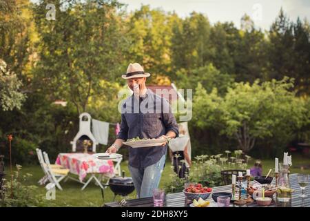 Fröhlicher Mann, der Teller am Esstisch im Hinterhof trägt Während Gartenparty Stockfoto