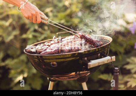 Zugeschnittene Hand der Frau, die Fleisch mit einer Servierzange grilt Hinterhof an sonnigen Tag Stockfoto