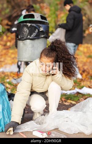 Junge freiwillige Helferin, die auf dem Smartphone spricht, während sie Müll sammelt Im Park Stockfoto