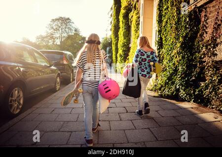 Rückansicht von Mädchen mit Skateboards, die auf dem Bürgersteig vorbeigehen Geparkte Autos an sonnigen Tagen Stockfoto