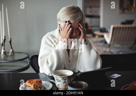 Besorgt ältere Frau mit Kopf in den Händen Blick auf digitale Tablet beim Sitzen am Esstisch zu Hause Stockfoto