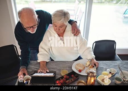 High-Angle-Ansicht des älteren Ehepaars, das digitale Tablets betrachtet Während Sie am Esstisch im Wohnzimmer sitzen Stockfoto