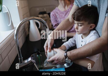 Mittelteil des Mannes, der Junge beim Abwaschen von Geschirr durch Frau unterstützt In der Küche Stockfoto