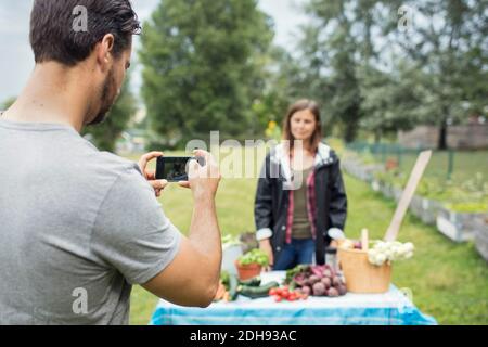 Mittelerwachsener Mann fotografiert Frau, die neben frisch geerntetem Gemüse steht Im Stadtgarten Stockfoto