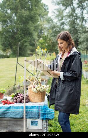 Mittlere Erwachsene Frau, die auf Pappe schreibt, während sie frisch dabeisteht Geerntetes Gemüse im städtischen Garten Stockfoto