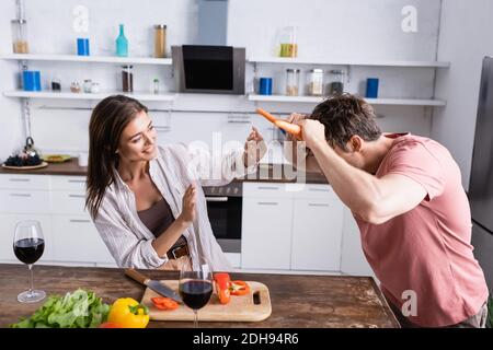 Mann hält Karotten neben dem Kopf neben lächelnder Frau, Gemüse und Wein auf dem Küchentisch Stockfoto