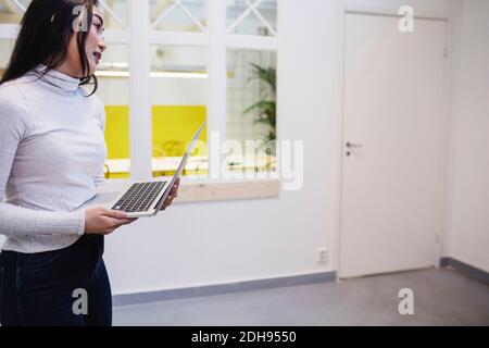 Junge Frau schaut weg, während sie Laptop in kreativem Büro hält Stockfoto