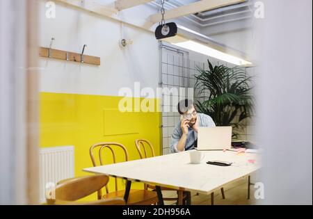 Junger Mann mit Smartphone und Laptop, während er sitzt Schreibtisch im Konferenzraum Stockfoto
