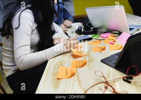 Mittelteil der jungen Frau, die auf Klebstoff-Notiz bei Holz schreibt Schreibtisch im Büro Stockfoto