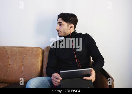 Der junge Mann schaut weg, während er Musik über ein digitales Tablet hört Auf dem Sofa im Büro Stockfoto