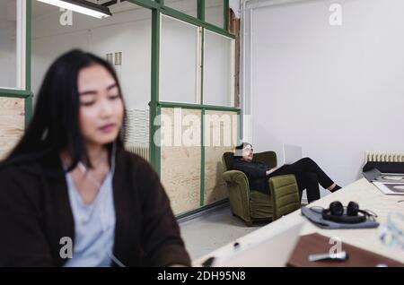 Junger Mann mit Laptop, während er sich auf einem Sessel mit Frau zurücklehnend Kollege im Vordergrund Stockfoto